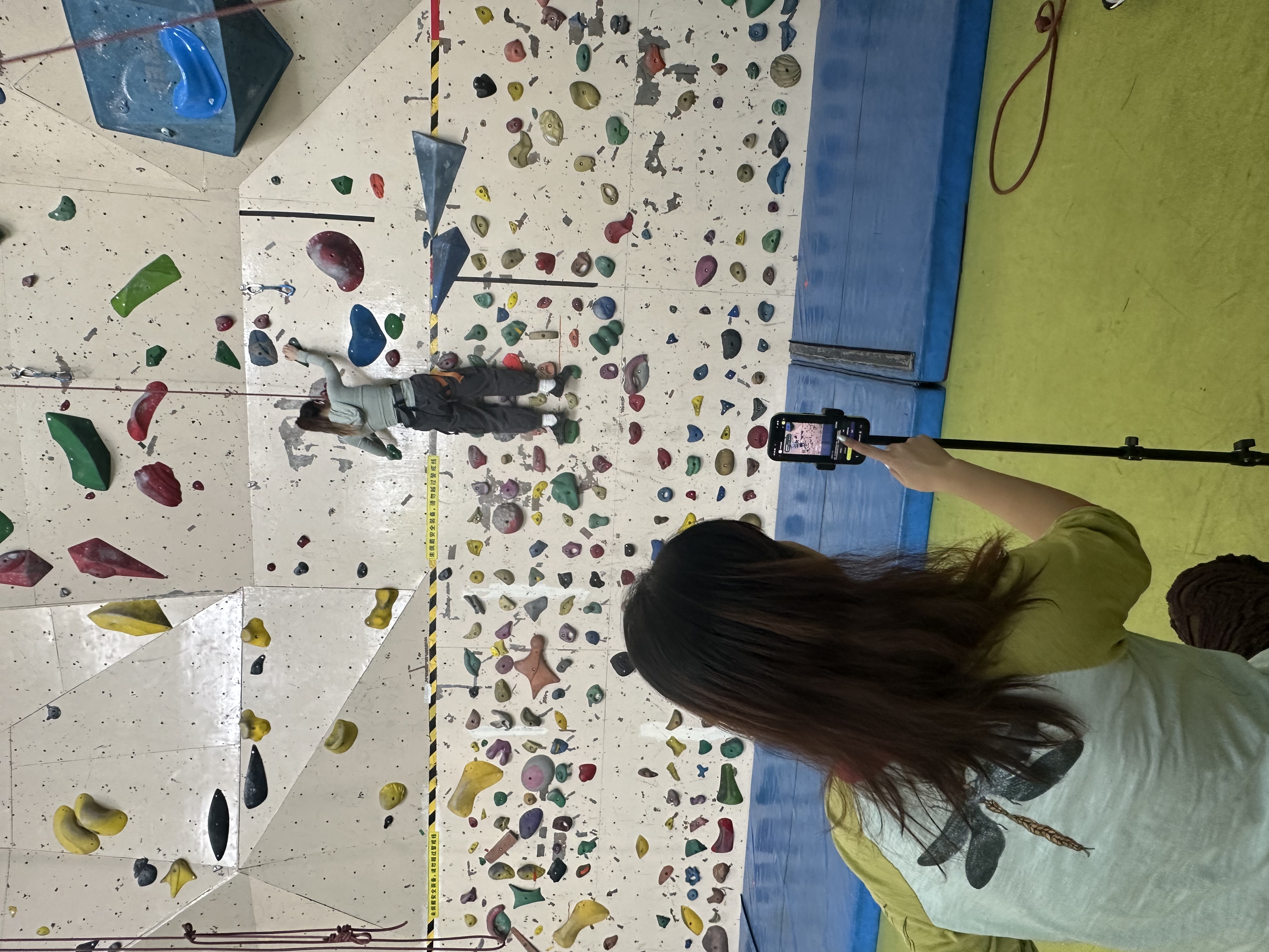 Final onsite testing setup showing the climber, guide, tripod-mounted phone, belayer support, and climbing wall context.